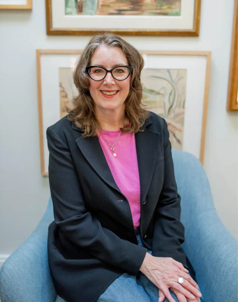 Woman with glasses and pink shirt under black blazer sits in blue chair.