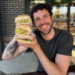 Man holding three stacked sandwiches in front of a brick building