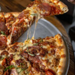 Slice of pizza with melted cheese, pepperoni, sausage, and basil